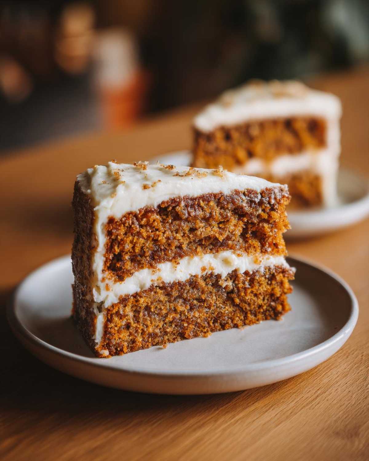 Two slices of moist pumpkin spice cake with creamy frosting on plates, sitting on a wooden table.