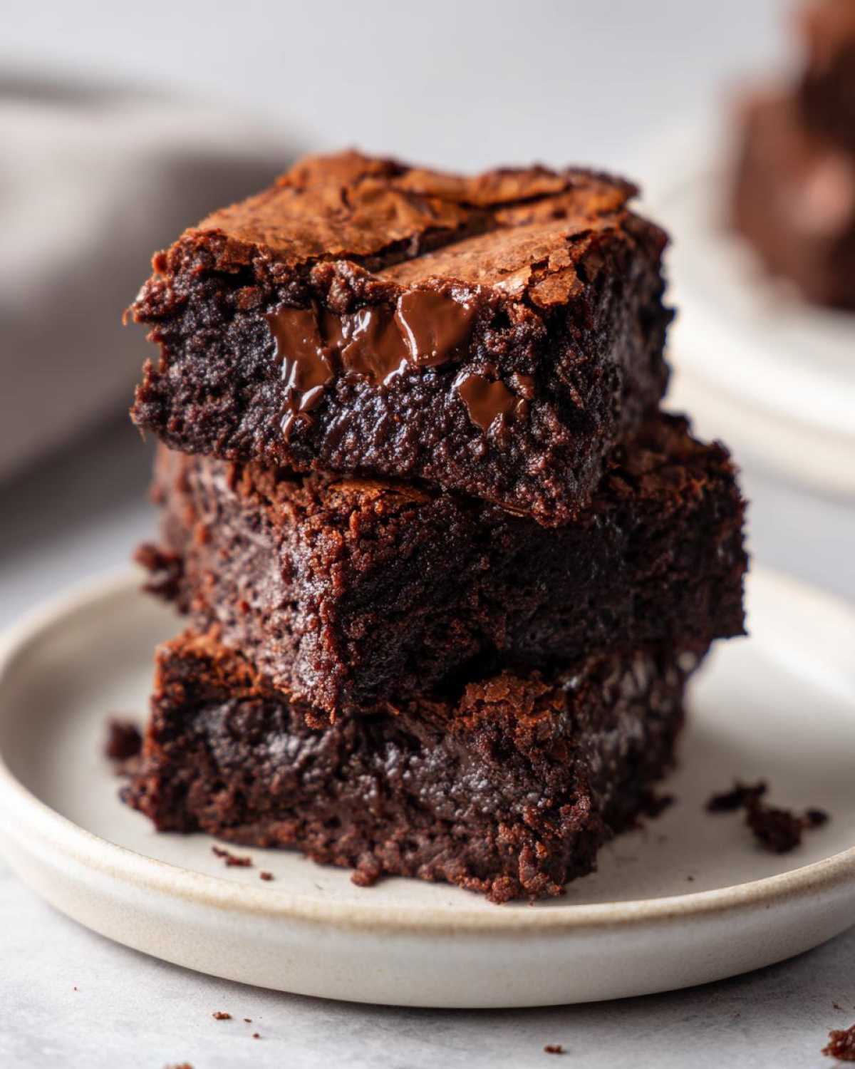 A close-up stack of three fudgy protein brownies on a white plate, showing melted chocolate chips and a rich texture.