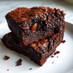 Two rich, dark chocolate protein brownies stacked on a white plate, showing a fudgy texture and visible chocolate chips.