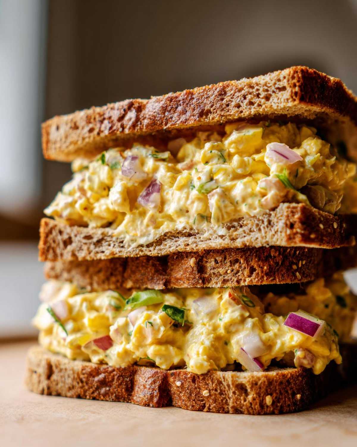 Close-up of a stacked tuna egg salad sandwich on whole wheat bread, showing creamy filling with red onion and herbs.