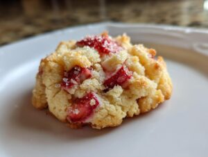 A single, freshly baked strawberry shortcake cookie on a white plate, topped with sugar crystals and visible strawberry pieces.