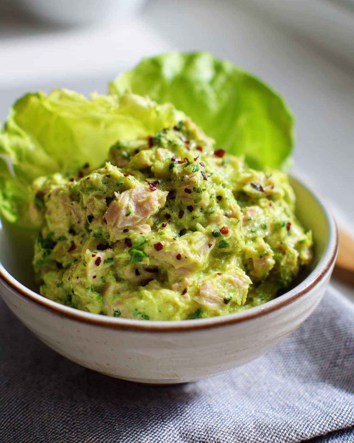 A close-up of a bowl filled with creamy tuna and avocado salad, garnished with red pepper flakes and fresh lettuce.