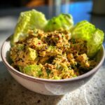 A close-up of a bowl filled with creamy tuna and avocado salad, garnished with red pepper flakes and fresh herbs, with lettuce leaves.
