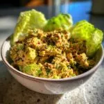 A close-up of a bowl filled with creamy tuna and avocado salad, garnished with red pepper flakes and fresh herbs, with lettuce leaves.