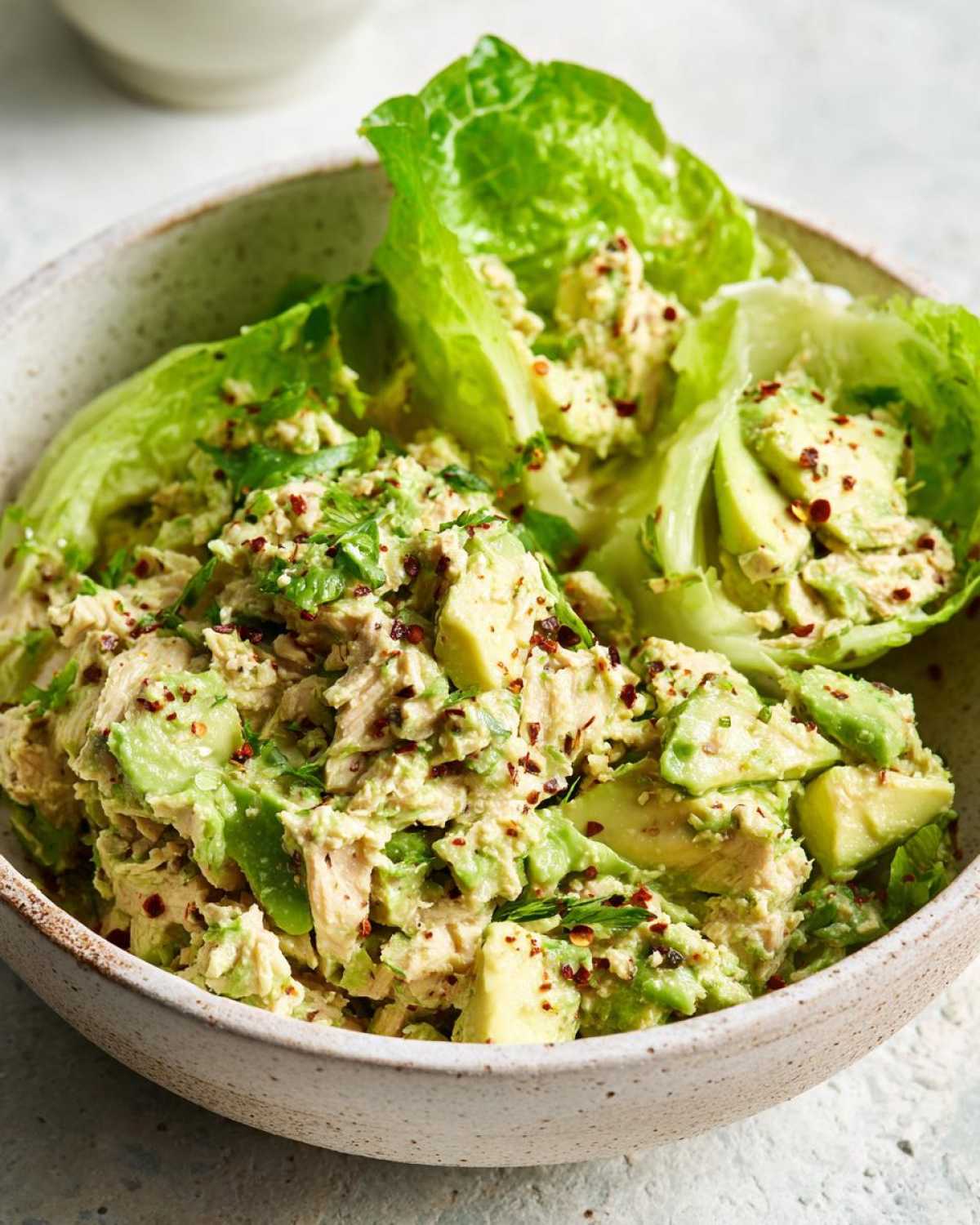 A close-up shot of a bowl filled with creamy tuna and avocado salad, garnished with red pepper flakes and fresh herbs.