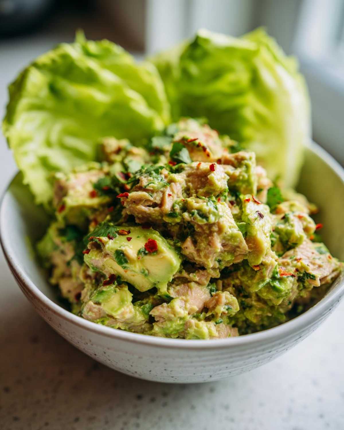 A close-up shot of a bowl filled with creamy tuna and avocado salad, garnished with red pepper flakes and cilantro, with lettuce in the background.