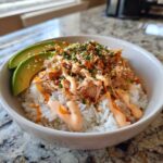A close-up of a white bowl filled with tuna and rice, topped with avocado slices, sauce, sesame seeds, and chives.