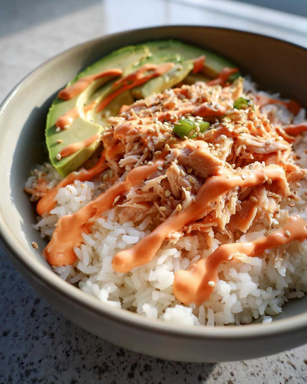 Close-up of a tuna and rice bowl topped with shredded tuna, avocado slices, creamy orange sauce, sesame seeds, and green onions.
