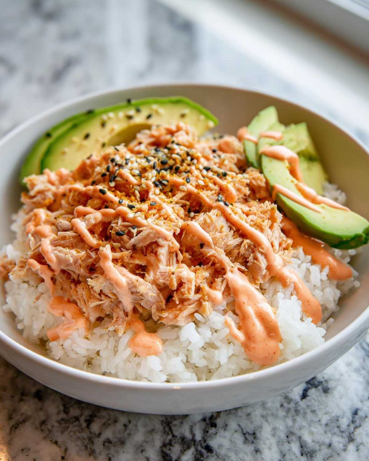 A close-up of a vibrant tuna and rice bowl topped with creamy tuna, avocado slices, and sesame seeds.
