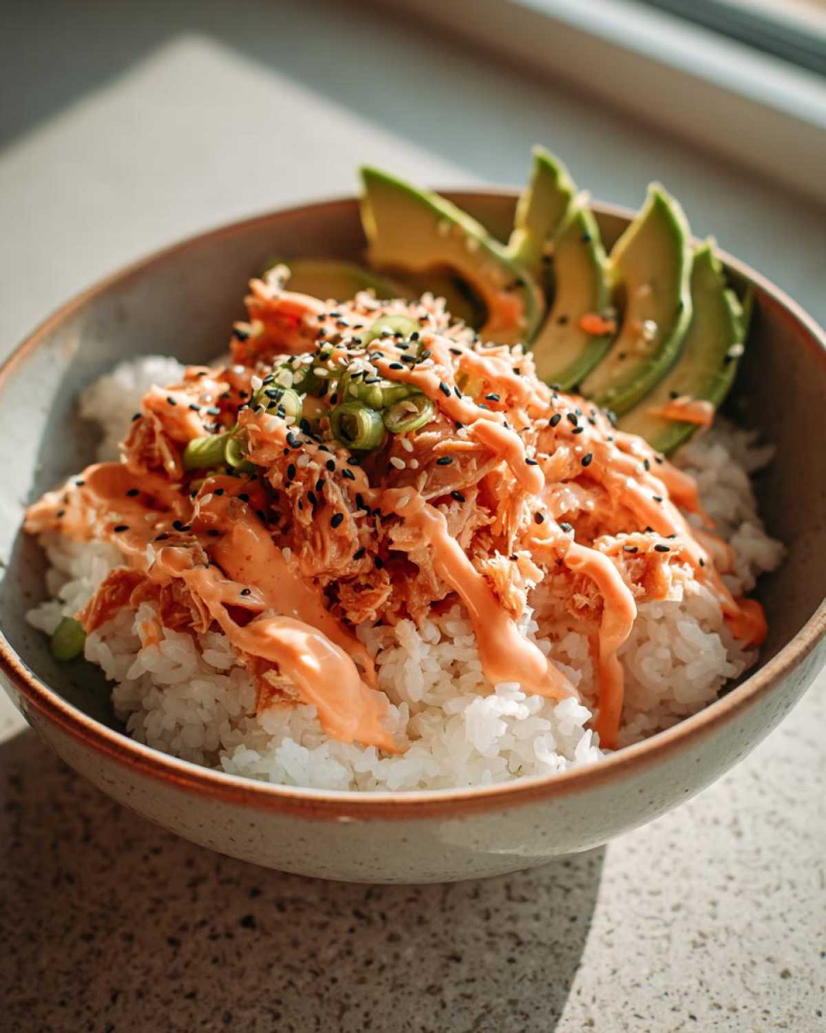 A vibrant tuna and rice bowl topped with spicy mayo, sesame seeds, green onions, and sliced avocado.