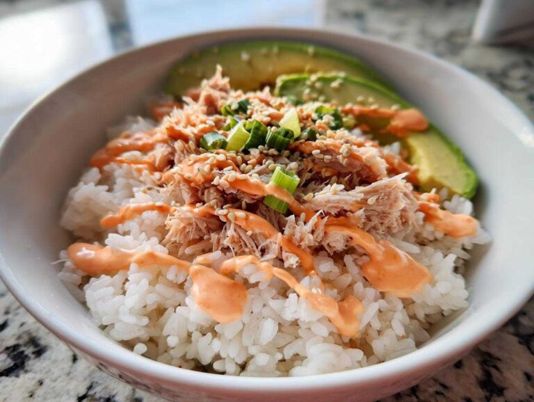 A close-up of a vibrant tuna and rice bowl topped with shredded tuna, creamy sauce, green onions, sesame seeds, and sliced avocado.