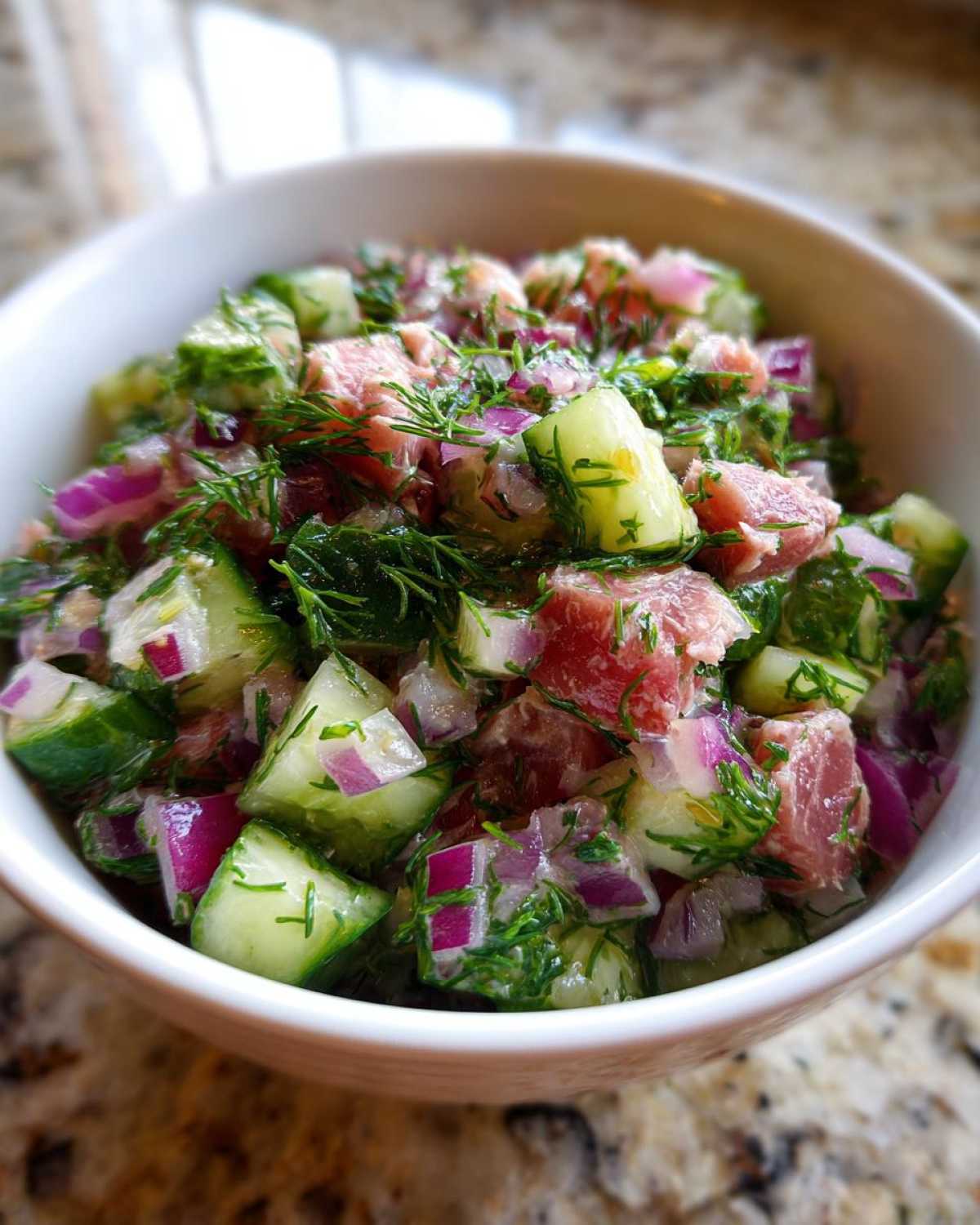 Close-up of a vibrant tuna cucumber salad in a white bowl, featuring diced tuna, cucumber, red onion, and fresh dill.