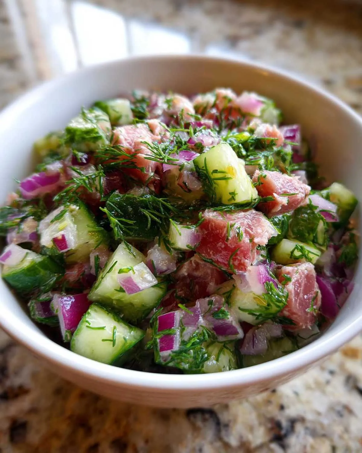 Close-up of a vibrant tuna cucumber salad in a white bowl, featuring diced tuna, cucumber, red onion, and fresh dill.
