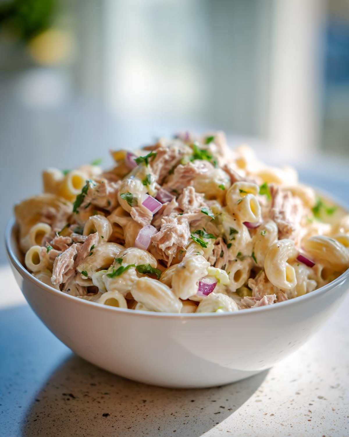 A close-up view of a white bowl filled with creamy tuna egg pasta salad, garnished with red onion and parsley.