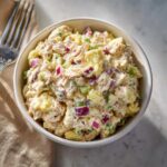 A close-up of a white bowl filled with creamy tuna egg salad, featuring chunks of tuna, yellow egg, red onion, and green celery.