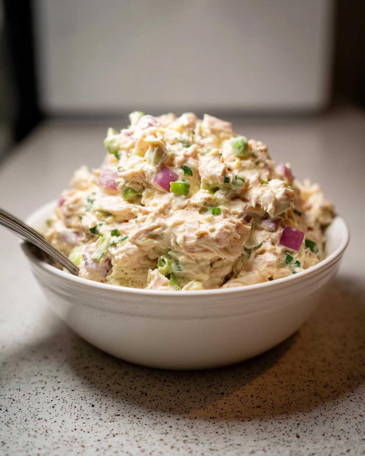 A close-up of a white bowl filled with creamy tuna egg salad, showing chunks of tuna, chopped red onion, and green herbs.