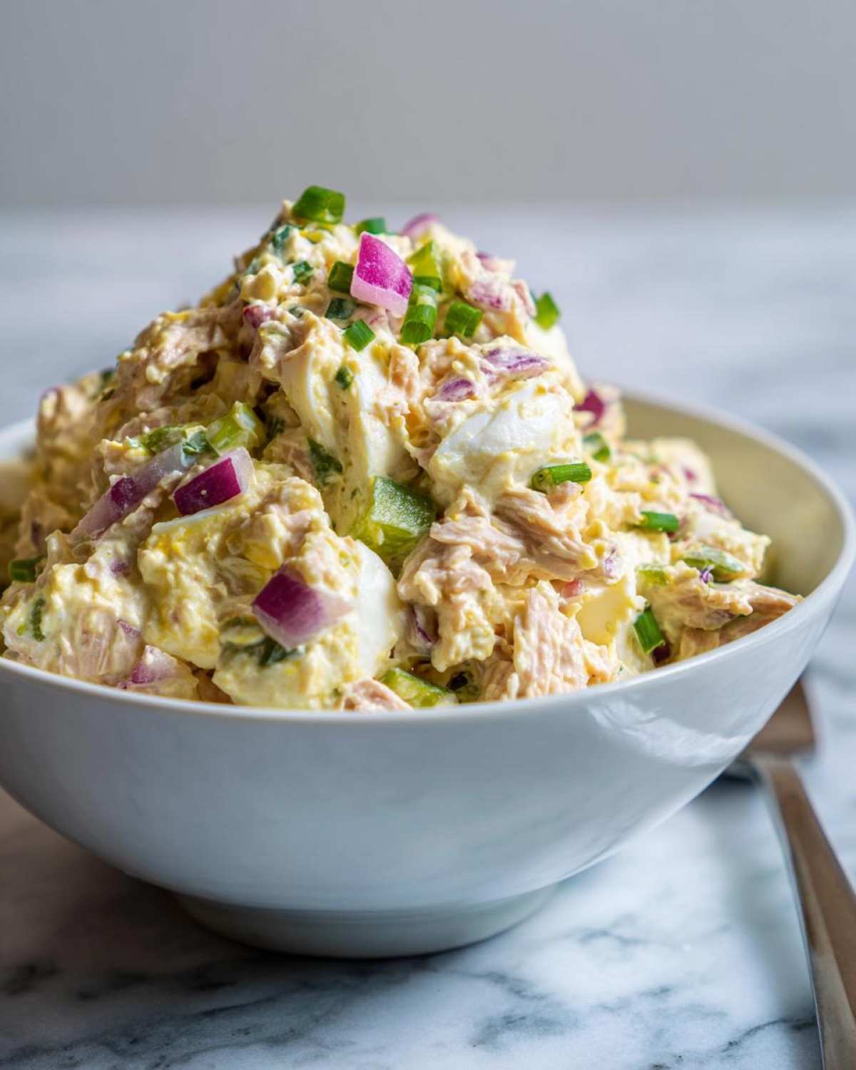 A close-up shot of a white bowl filled with creamy tuna egg salad, garnished with red onion and green onions.