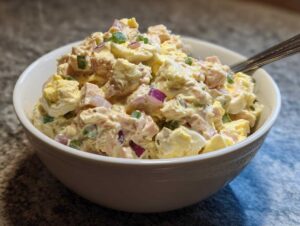 A close-up of a white bowl filled with creamy tuna egg salad, showing chunks of tuna, hard-boiled eggs, red onion, and green onion.