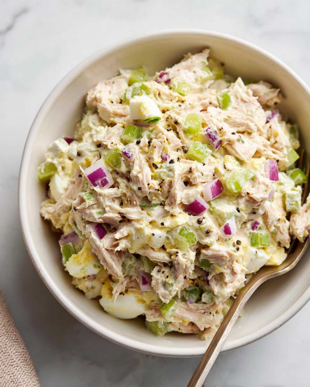 A close-up shot of a bowl filled with creamy tuna egg salad, featuring visible chunks of tuna, hard-boiled eggs, celery, and red onion, sprinkled with black pepper.