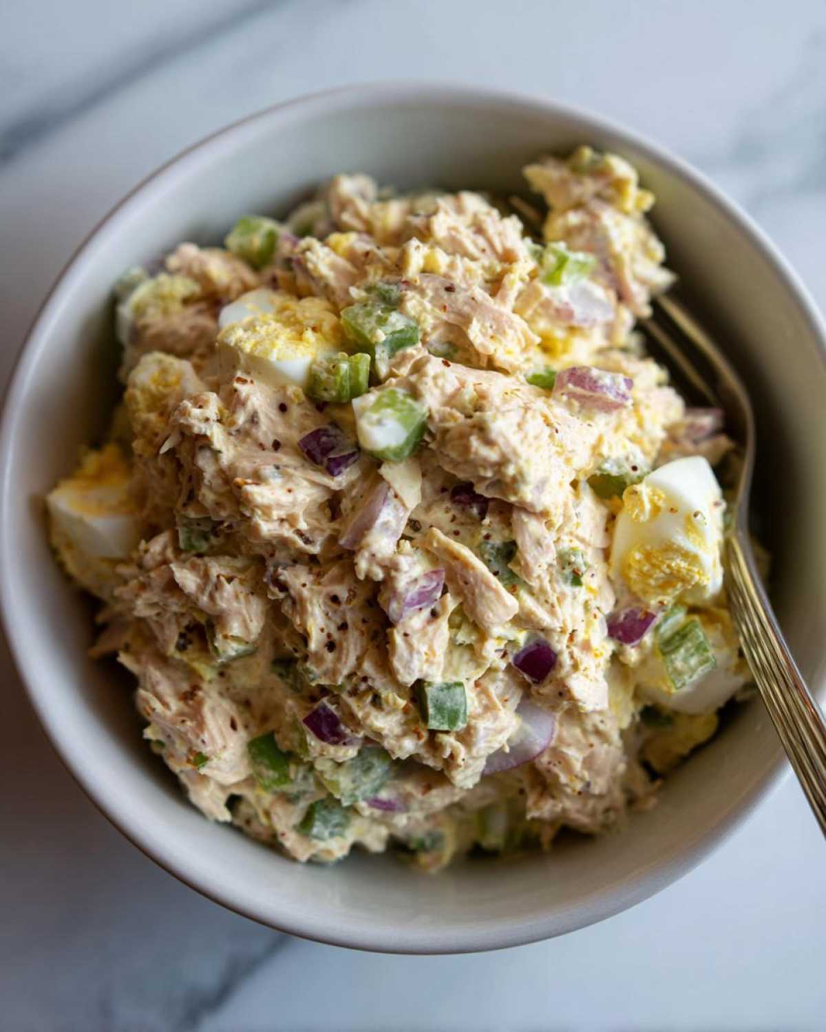 A close-up overhead shot of a bowl filled with creamy tuna egg salad, showing chunks of tuna, diced hard-boiled eggs, red onion, and celery.