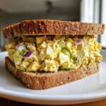 A close-up of a delicious tuna egg salad sandwich on whole wheat bread, showing chunks of egg, tuna, red onion, and green bell pepper.