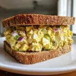 A close-up of a delicious tuna egg salad sandwich on whole wheat bread, showing chunks of egg, tuna, red onion, and green bell pepper.