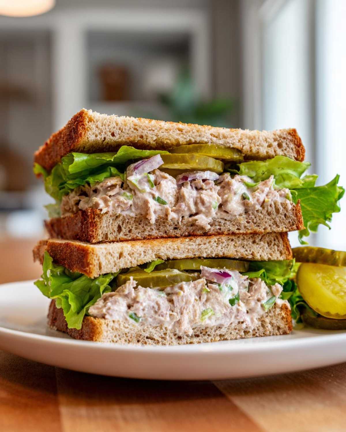 A perfectly assembled tuna fish sandwich on whole wheat bread with lettuce, red onion, and pickles, served on a white plate.