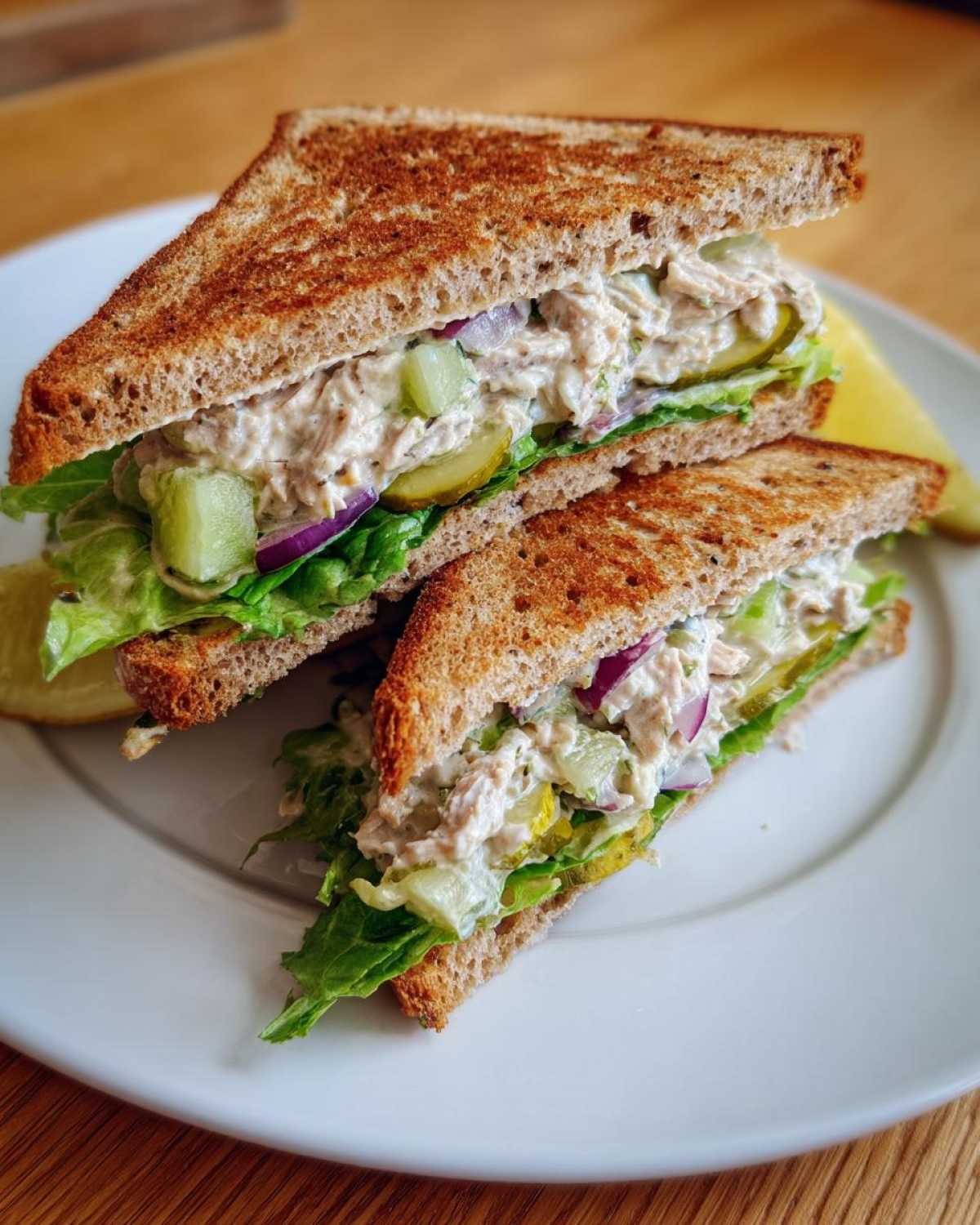 A delicious tuna fish sandwich cut in half, showcasing creamy tuna salad, lettuce, red onion, and pickles on toasted whole wheat bread.