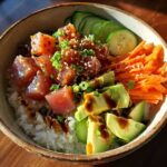 A close-up of a colorful tuna rice bowl featuring diced tuna, sliced avocado, shredded carrots, and cucumber.