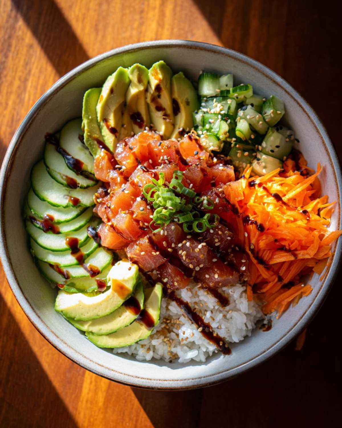 A colorful tuna rice bowl featuring diced tuna, sliced avocado, cucumber, shredded carrots, and a drizzle of sauce.