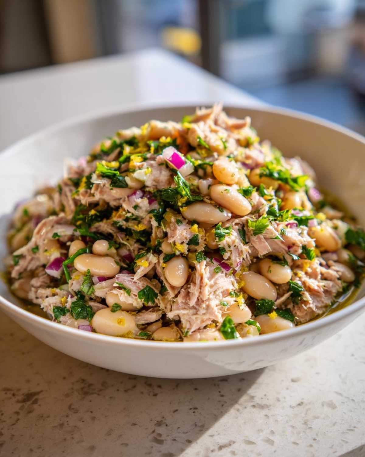 A close-up shot of a vibrant tuna white bean salad in a white bowl, garnished with red onion, parsley, and lemon zest.