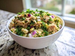 A close-up of a white bowl filled with a vibrant tuna white bean salad, garnished with red onion and parsley.