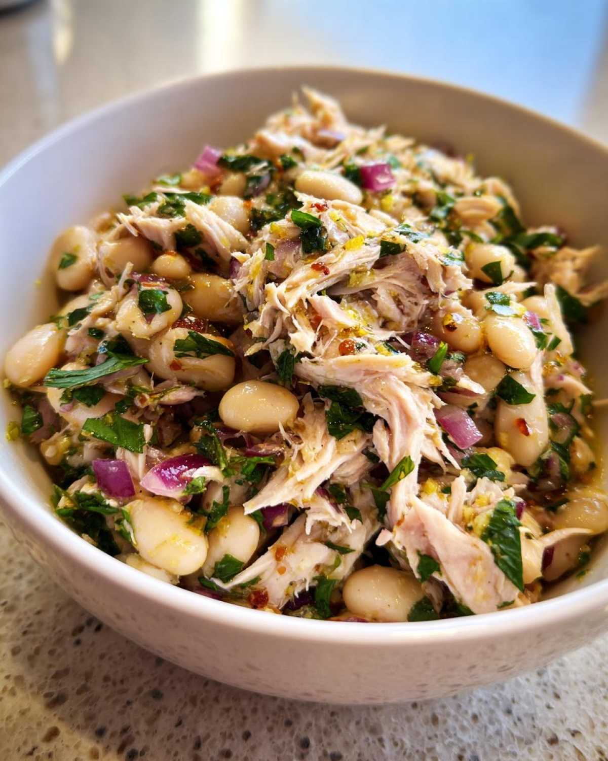 A close-up shot of a white bowl filled with a vibrant tuna white bean salad, showing white beans, shredded tuna, red onion, and fresh herbs.