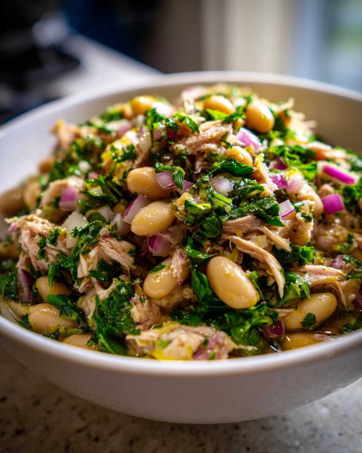 Close-up shot of a bowl filled with tuna white bean salad, showing white beans, flaked tuna, chopped red onions, and green herbs.
