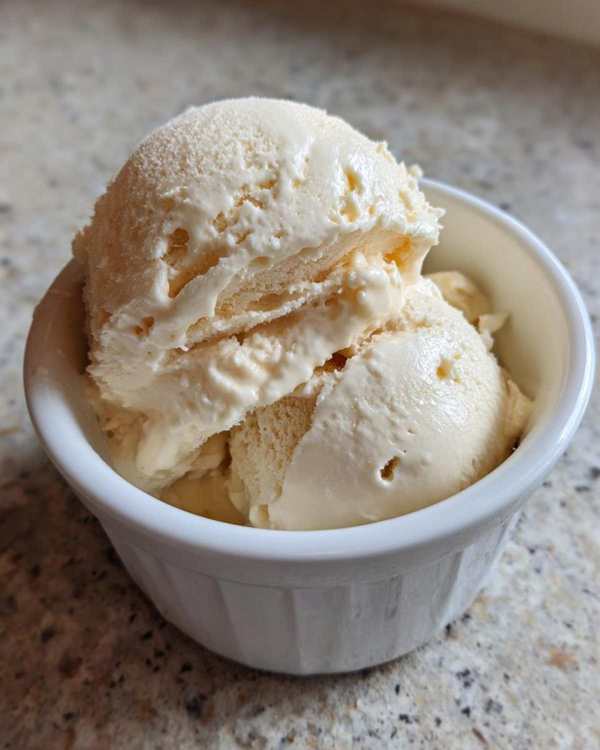 Close-up of several scoops of creamy, light-colored protein ice cream in a white bowl on a speckled countertop.