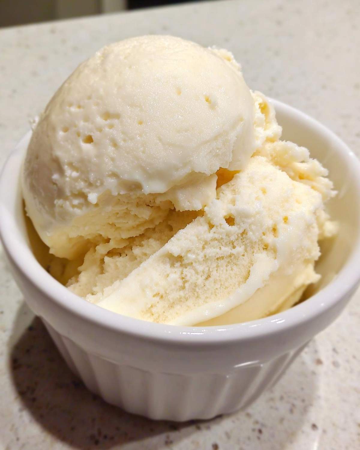 Close-up of two scoops of creamy, light yellow protein ice cream in a small white ceramic bowl on a speckled countertop.