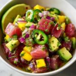 Close-up of fresh tuna ceviche in a white bowl, featuring diced tuna, mango, avocado, red onion, and sliced jalapeños, garnished with cilantro and a lime wedge.