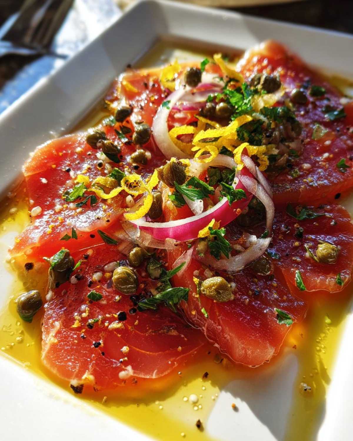 Close-up of fresh tuna crudo slices on a white plate, garnished with capers, red onion, lemon zest, and herbs.