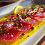 Close-up of a vibrant tuna crudo dish with thin slices of red tuna, capers, red onion, parsley, and lemon zest in olive oil, garnished with a lemon slice.