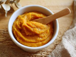 A white bowl filled with smooth, vibrant orange pumpkin puree, with a wooden spoon resting inside.