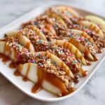 Close-up of apple slices arranged on a white platter, drizzled with caramel and topped with crunchy toppings and colorful sprinkles for a caramel apple bar.