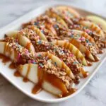 Close-up of apple slices arranged on a white platter, drizzled with caramel and topped with crunchy toppings and colorful sprinkles for a caramel apple bar.