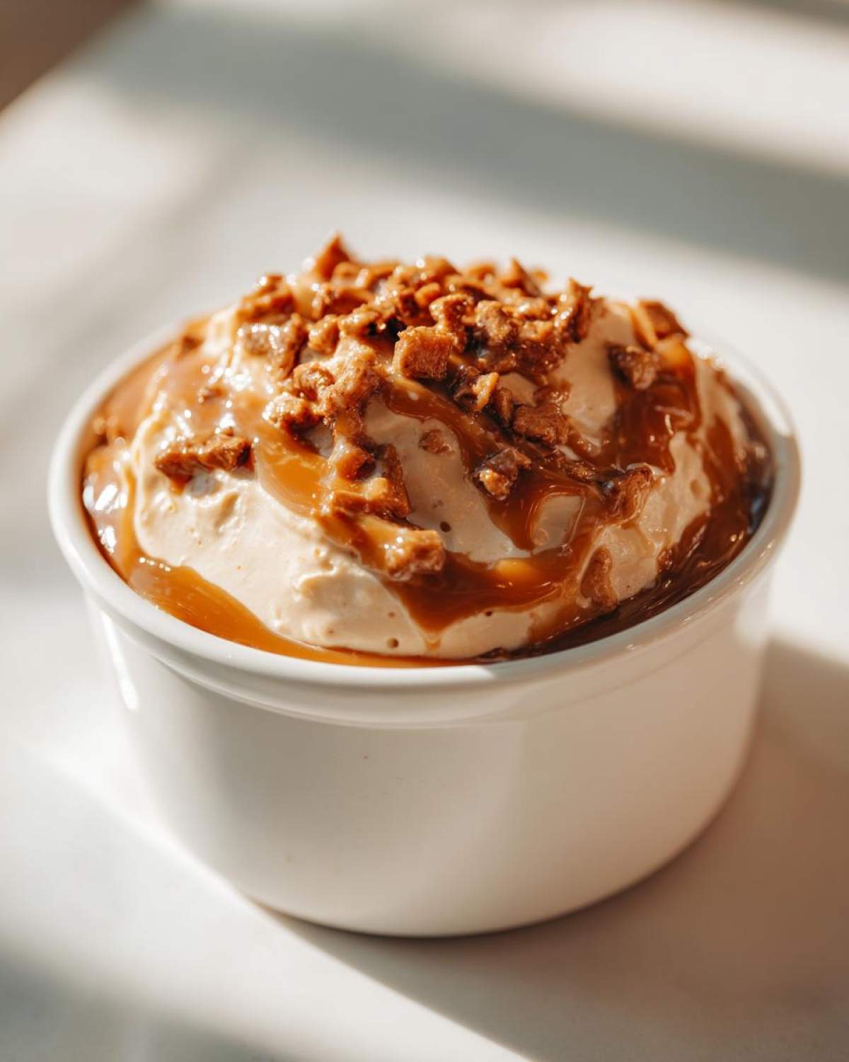 A close-up of a creamy caramel apple dip topped with caramel sauce and crunchy toffee pieces in a white ramekin.
