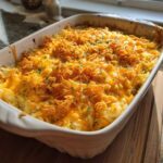 A close-up of a cheesy hashbrown casserole in a white baking dish, topped with melted cheddar cheese and crunchy cornflakes.