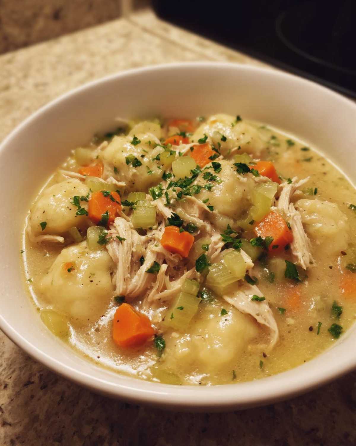 A close-up of a bowl of chicken and dumpling soup, featuring shredded chicken, carrots, celery, and fluffy dumplings, garnished with parsley.