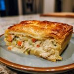 A close-up of a slice of chicken pot pie with a flaky golden crust and creamy chicken and vegetable filling.