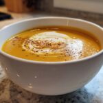 A close-up of a bowl of creamy pumpkin soup, topped with a swirl of cream and a sprinkle of spice.