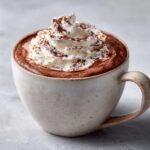 A close-up of a creamy mug of crockpot hot chocolate topped with whipped cream and a dusting of cocoa powder.