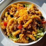 A vibrant bowl of Dorito taco salad pasta, featuring pasta, ground beef, lettuce, tomatoes, olives, and shredded cheese.