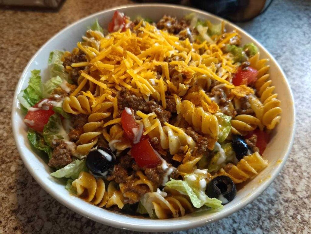 A colorful bowl of Dorito taco salad pasta with lettuce, tomatoes, olives, ground beef, cheese, and crushed Doritos.
